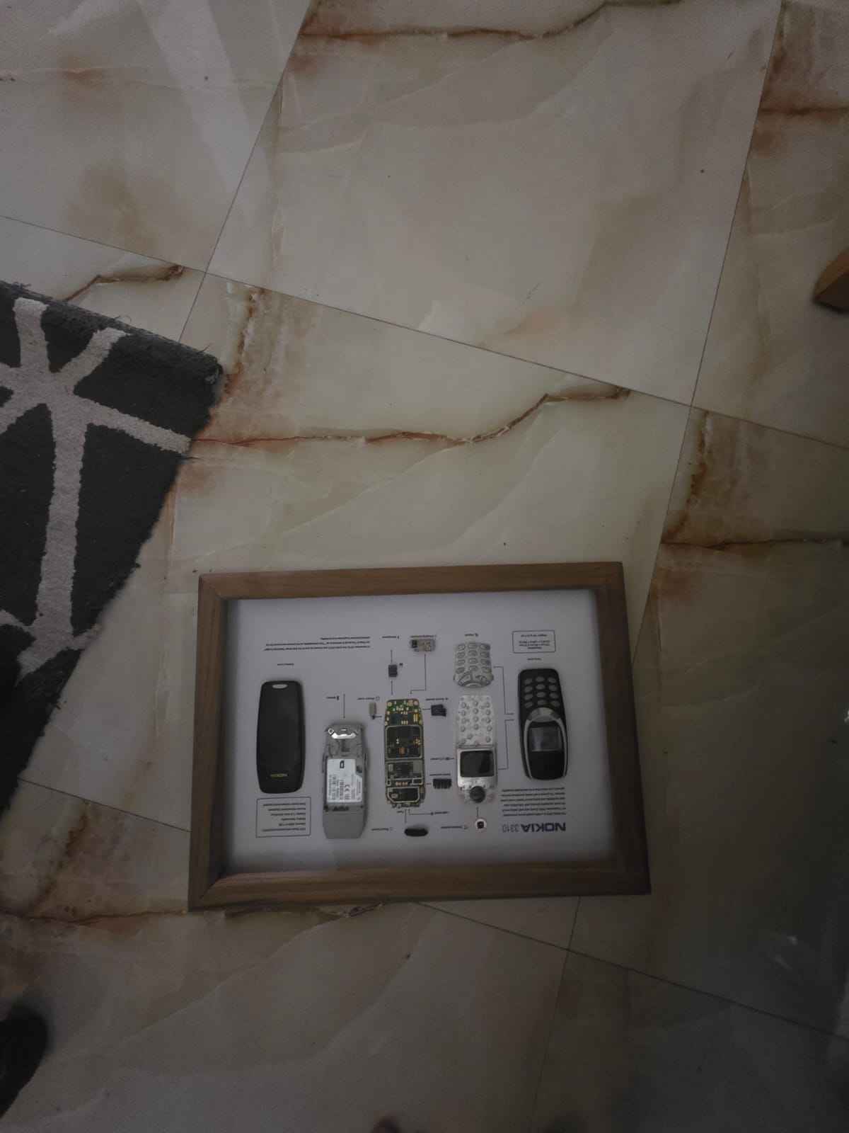 A framed teardown display of a disassembled Nokia 3310 mobile phone on a wooden background.